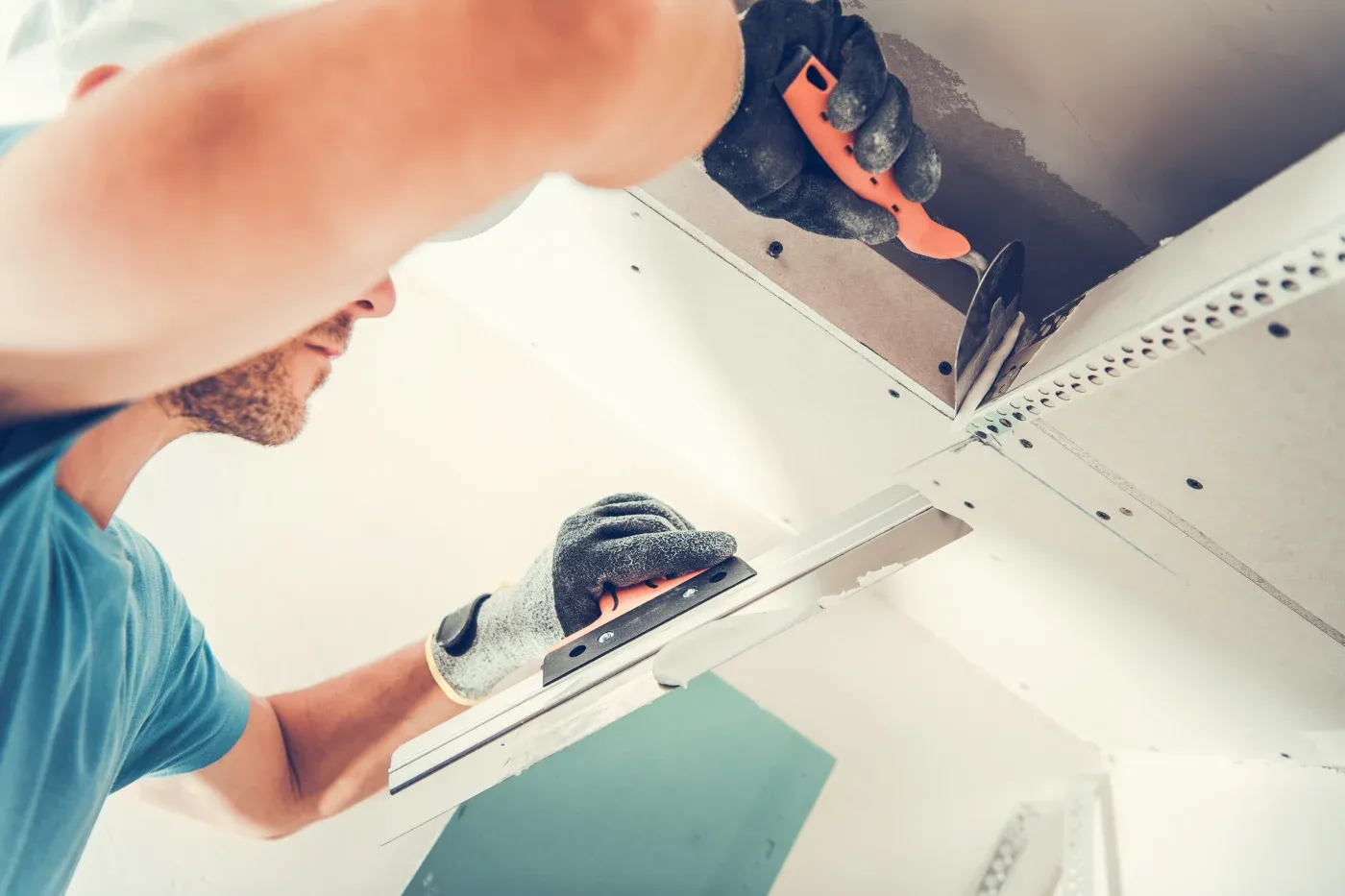 Worker in blue shirt and gloves using tools to install drywall on a ceiling, concentrating on smoothing and aligning the panels.