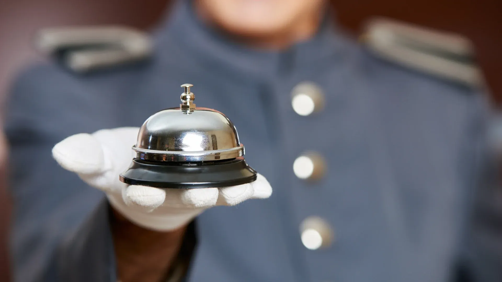 A hotel bellhop in a blue uniform and white gloves presents a shiny service bell on a tray. The image conveys professionalism and readiness to assist.