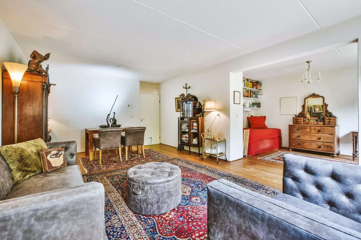 Sophisticated living room with gray sofas, red Persian rug, and a cozy work desk. Adjacent space has a red daybed and wooden dresser, conveying warmth.