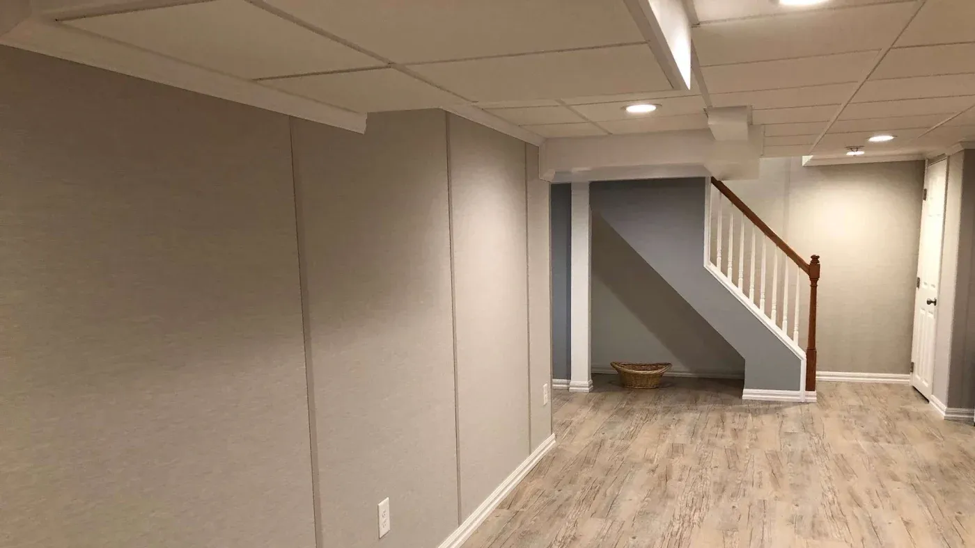 A modern basement features light gray walls, recessed lighting, and wood-like flooring. A staircase with a white railing adds depth. A wicker basket sits nearby, creating a cozy, minimalist space.