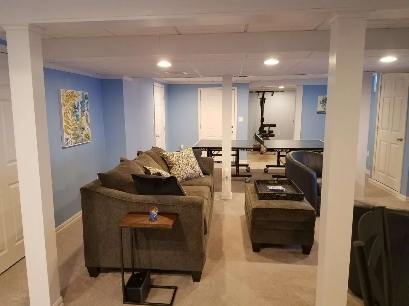 Basement living area with blue walls featuring a gray sectional sofa, a ping pong table in the background, and colorful wall art, creating a cozy and recreational atmosphere.