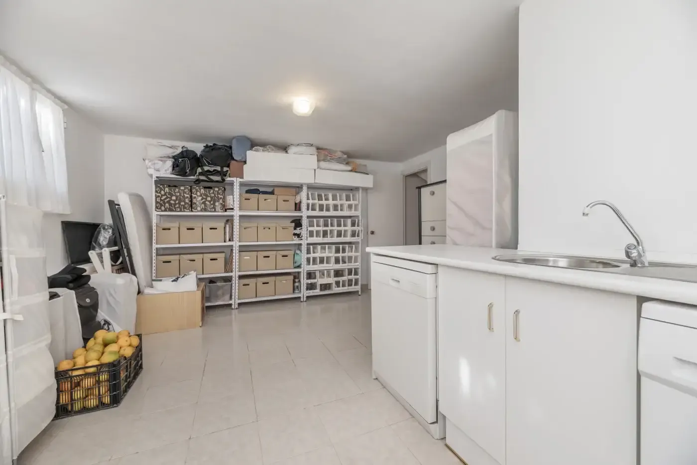 Laundry room with white cabinets and a sink on the right. Shelves with storage boxes and a crate of lemons on the left. Bright, organized space.