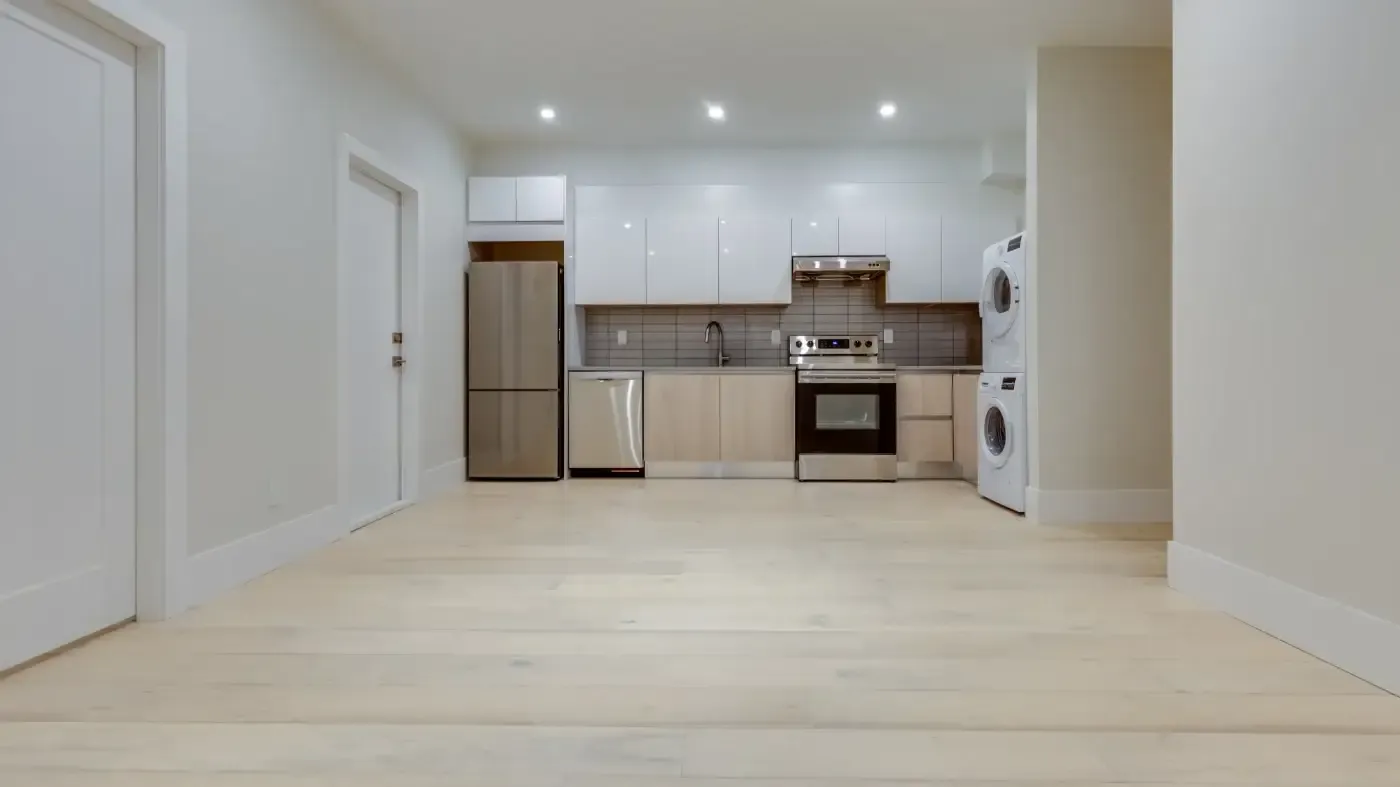 Modern, minimal kitchen with light wood flooring, white cabinets, stainless steel appliances, and a stacked washer-dryer. Bright, clean atmosphere.