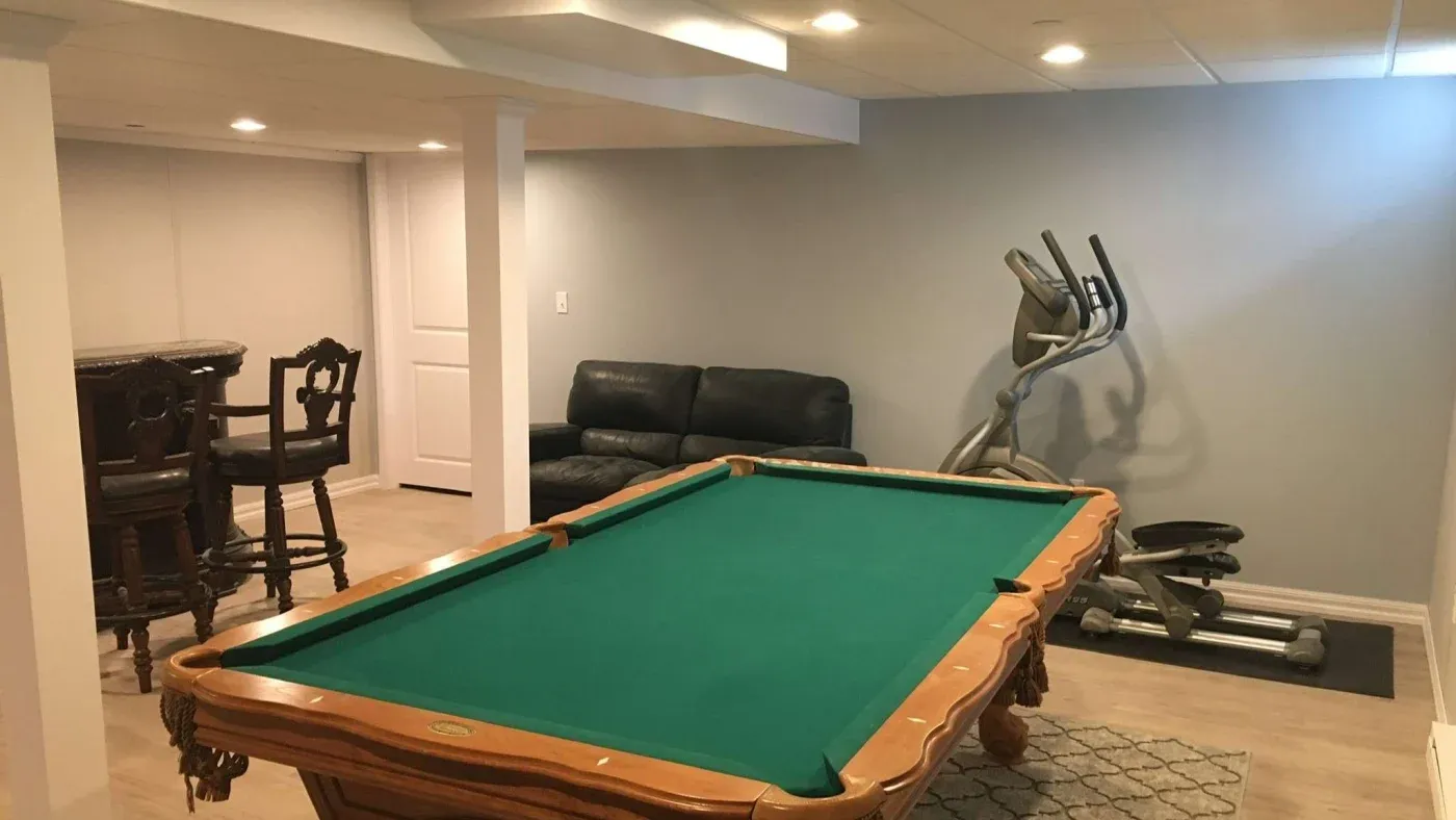 Basement rec room with a green pool table at center, flanked by a black leather sofa and elliptical machine. Bar area with wooden stools in the corner.