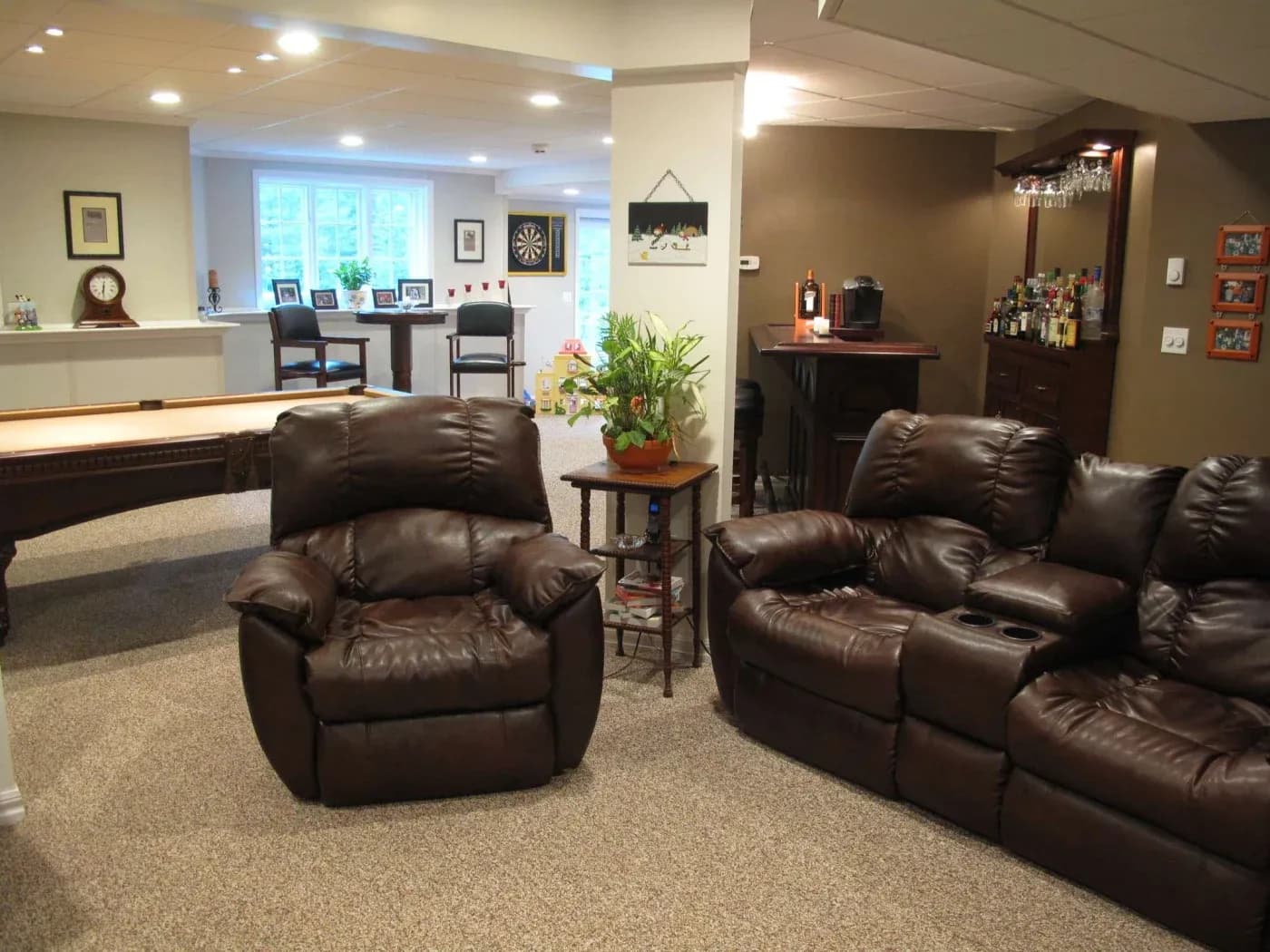 Cozy basement lounge with leather recliners, dartboard, and bar area. Soft lighting, plush carpet, and houseplant add warmth to the entertainment space.