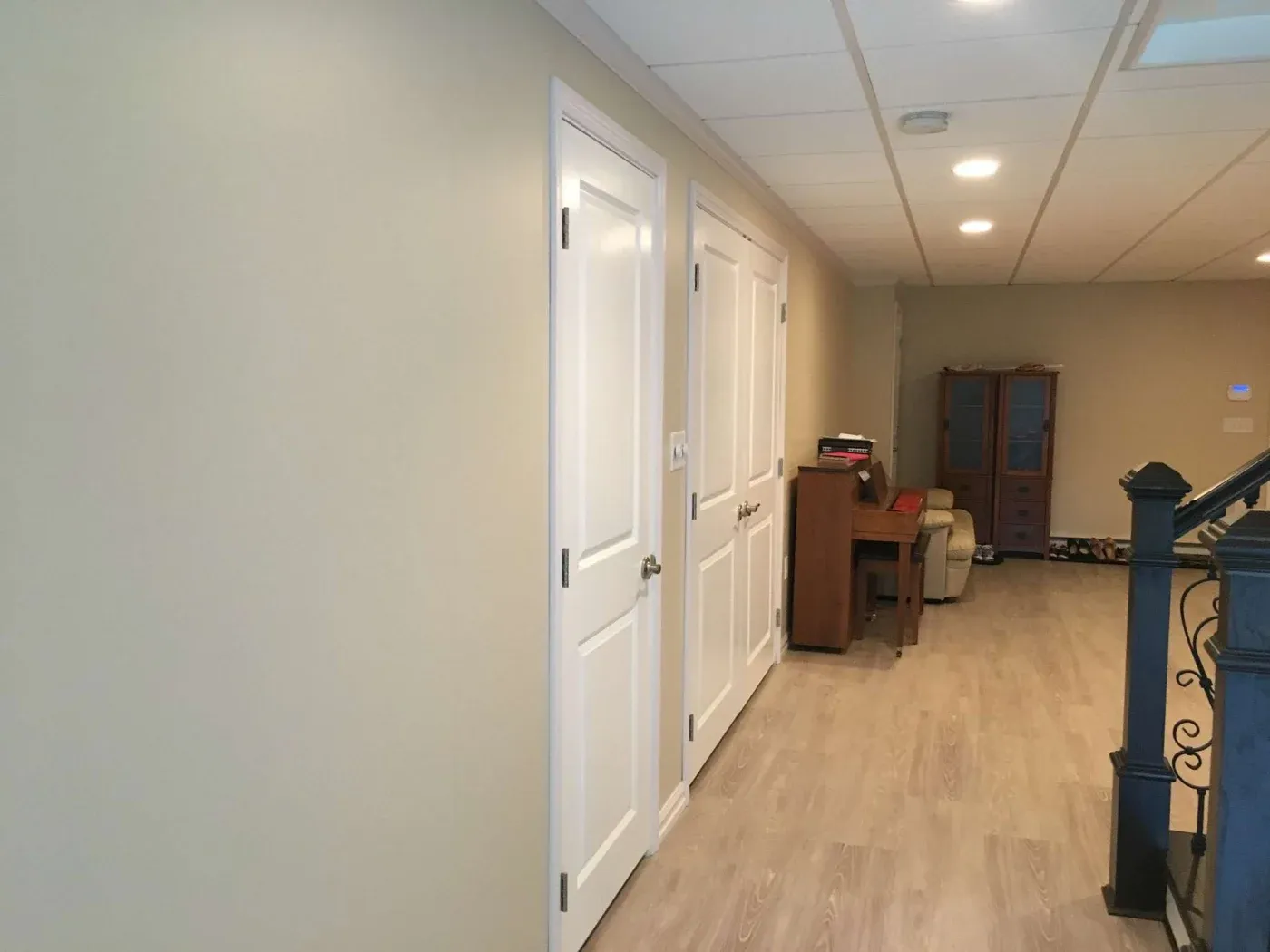 Spacious basement hallway with light beige walls, wood flooring, white doors on the left, and a seating area with a desk and cabinet at the end. Cozy and organized.