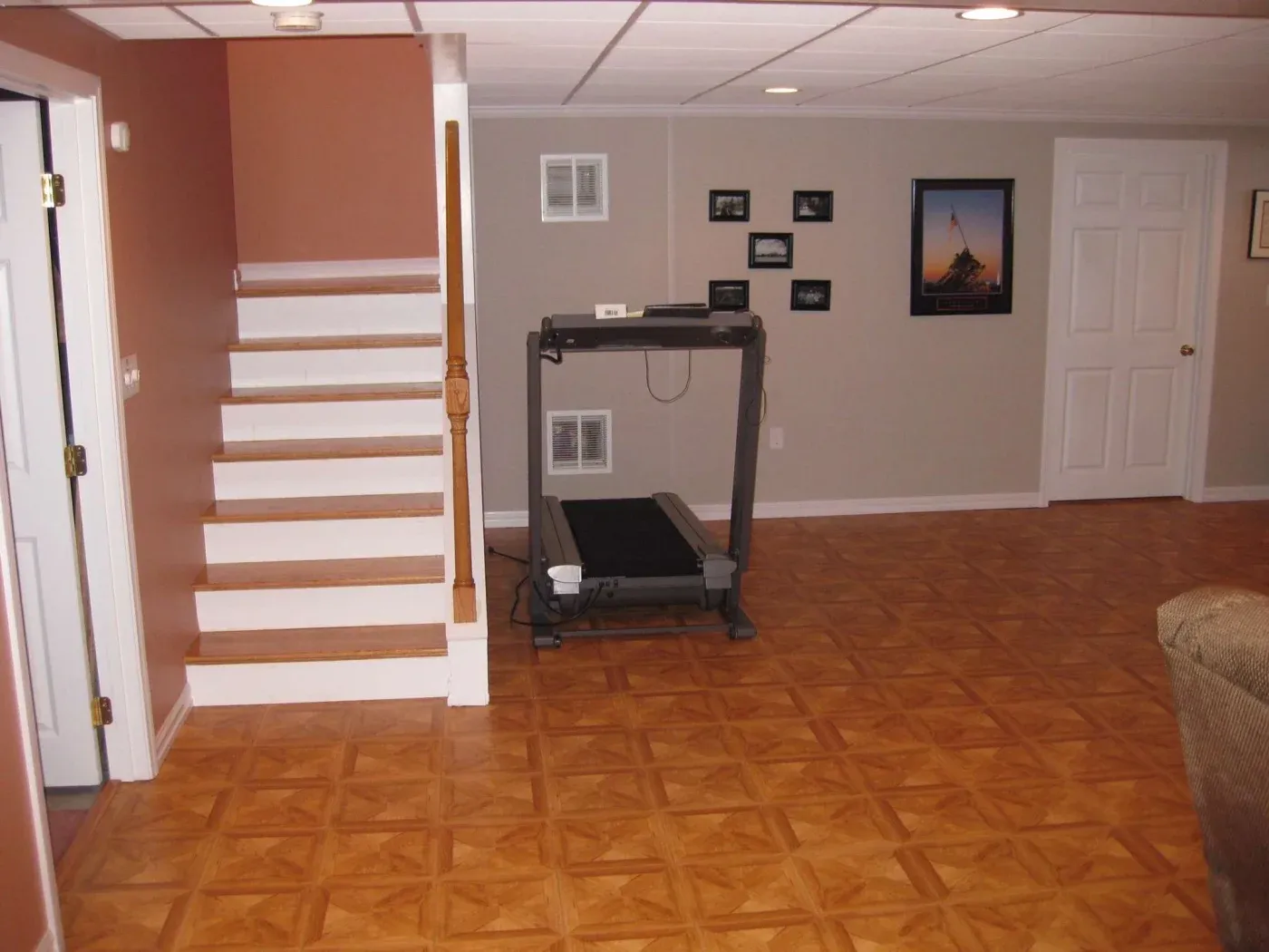 A tidy basement with parquet flooring, beige walls, and a white ceiling. A treadmill stands against the wall near framed photos and a closed door.