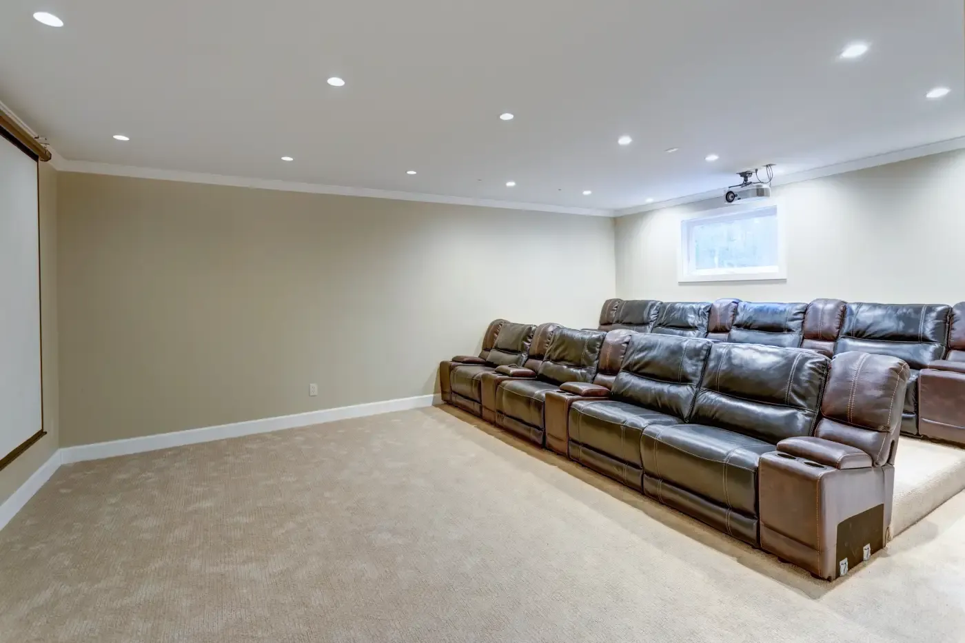 A home theater with brown leather recliners in two rows, beige walls, and a large screen. The room feels cozy and modern, with dim ceiling lights.