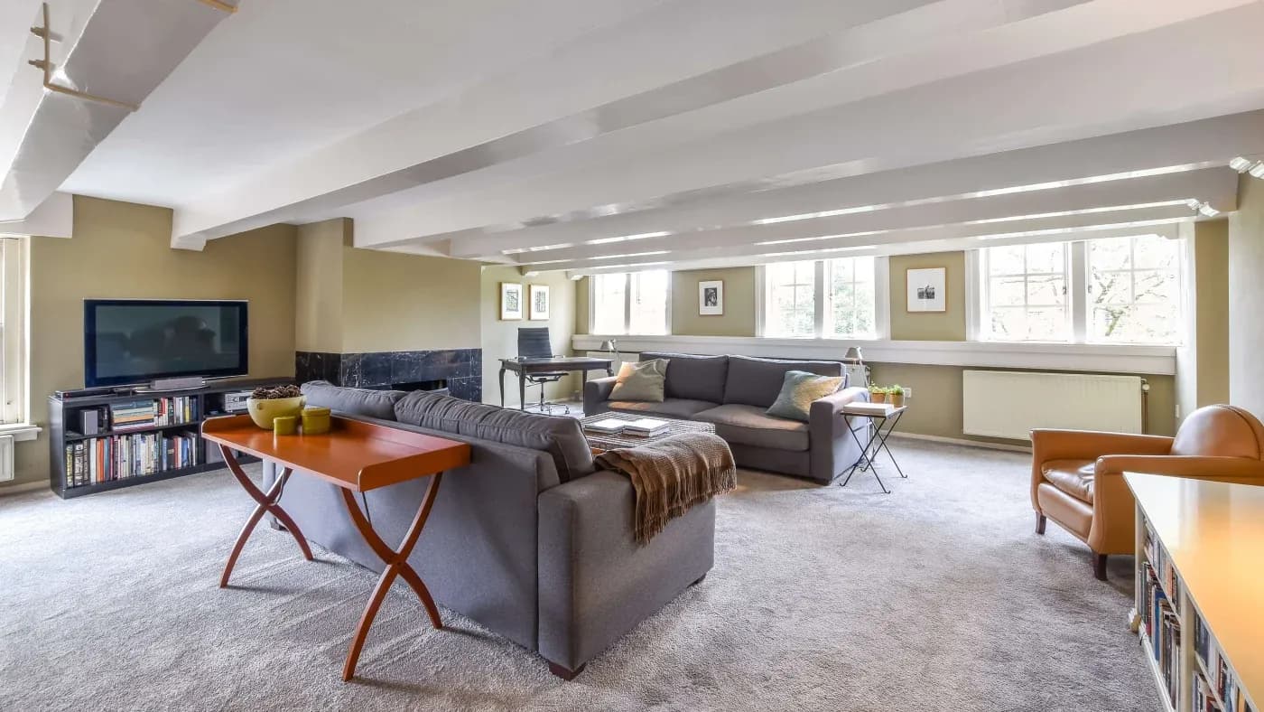 Spacious living room with gray sofas, a TV, bookshelves, and a wooden table with green vases. Large windows fill the room with natural light. Cozy ambiance.