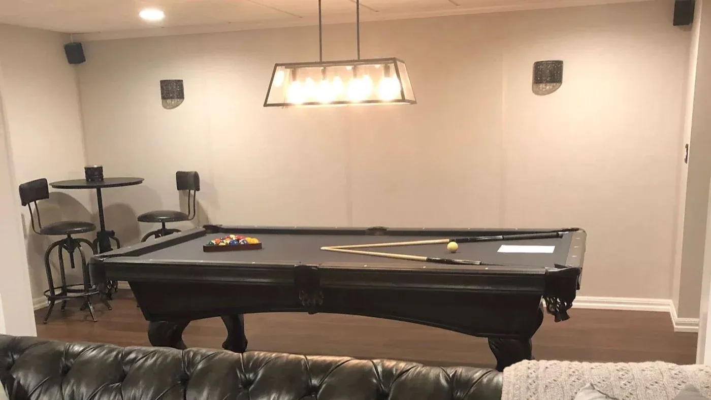 A cozy game room with a billiard table beneath a modern, rectangular light fixture. Bar stools and table in the corner add to the relaxed ambiance.