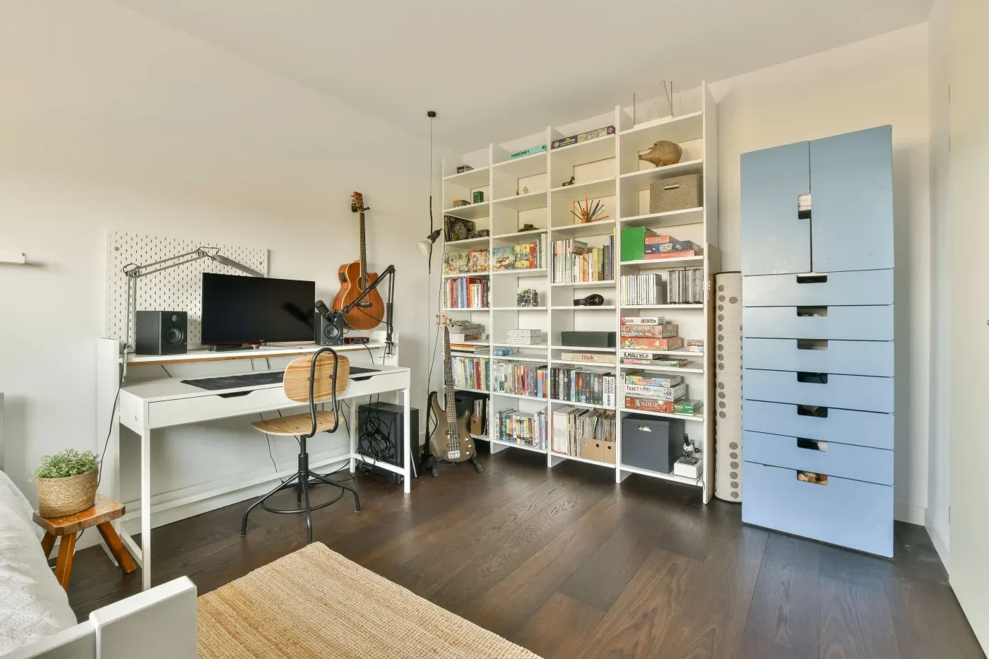 Modern home office with a white desk, computer, and chair. Guitars hang on the wall. A large bookshelf filled with books, decor, and games adds a cozy feel. A tall blue cabinet stands to the right.