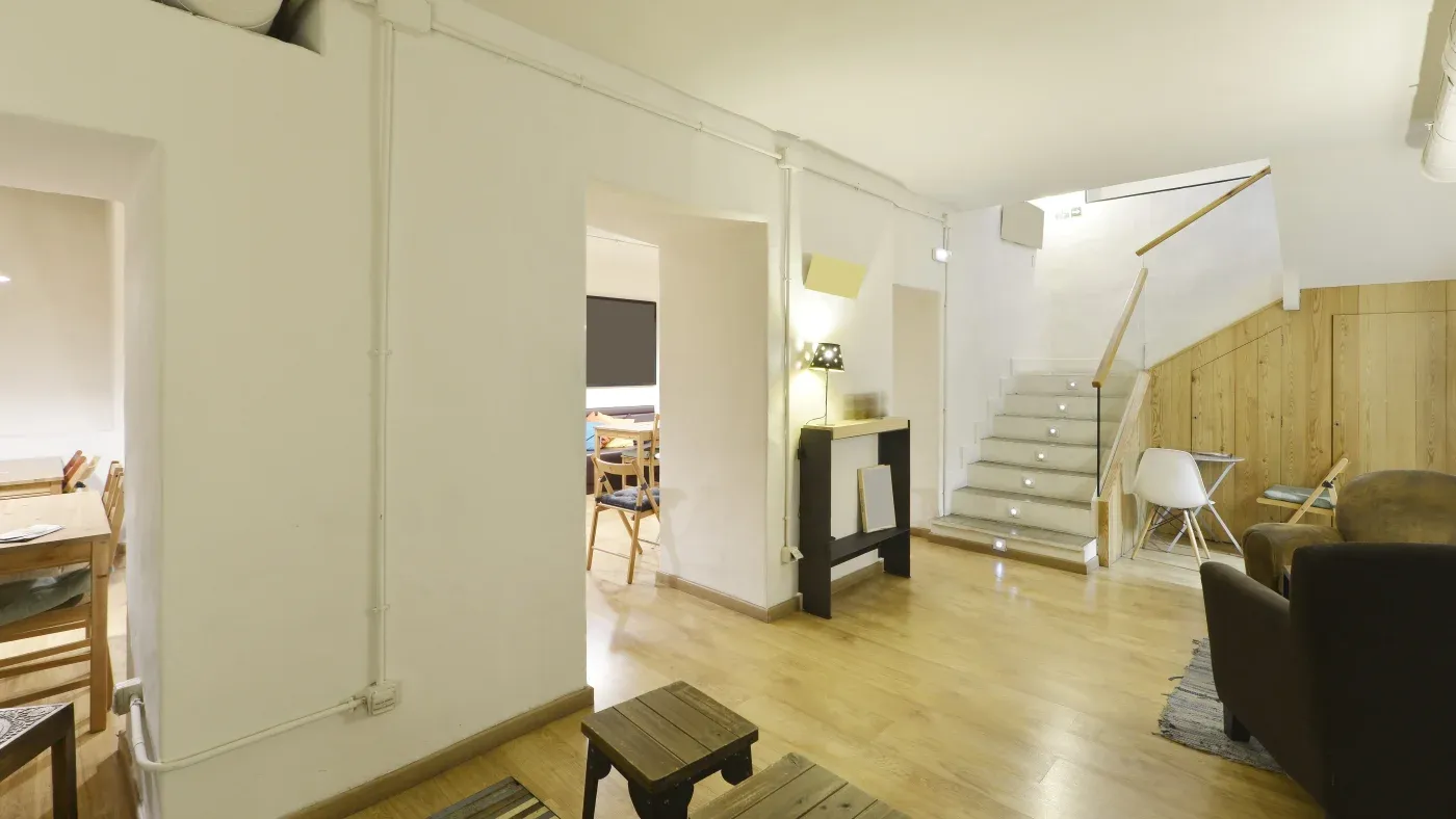 Modern living area with wooden floors, white walls, and stairs leading up. A dining table is visible through an archway, creating a cozy, inviting ambiance.