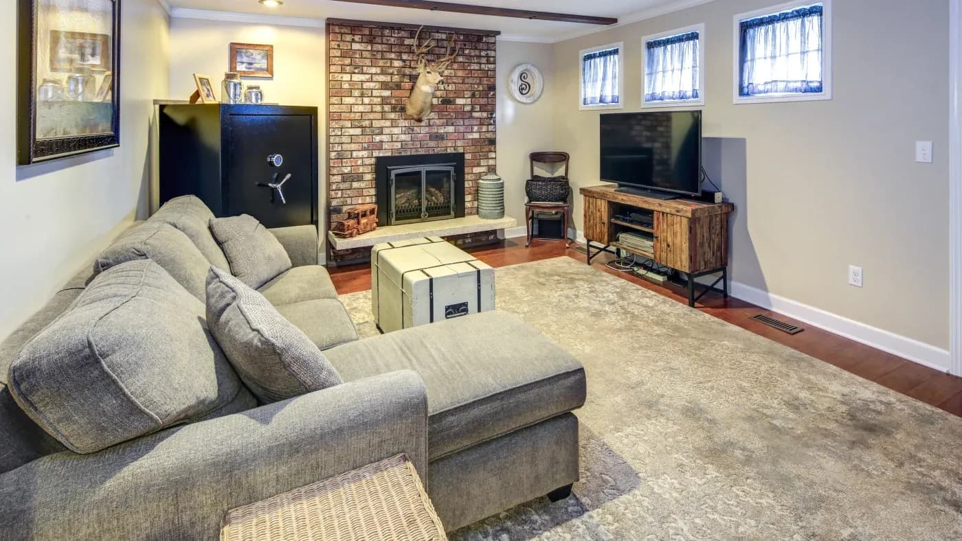 Cozy living room with a gray sectional sofa facing a brick fireplace. Decor includes a mounted deer head, TV, large safe, and soft lighting.