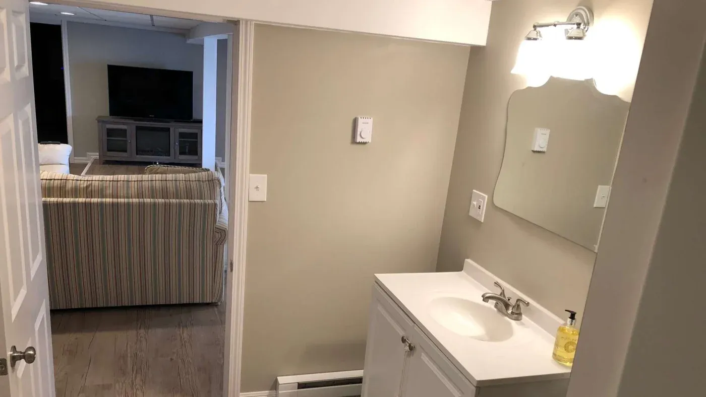 Small bathroom with a white vanity and mirror, lit by a wall-mounted light. Beyond, a cozy living room with a striped couch faces a large TV.