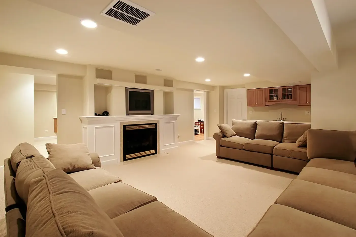 Spacious basement living room with beige walls and carpet. Features a large sectional sofa, TV above a fireplace, and a small kitchenette with wooden cabinets.