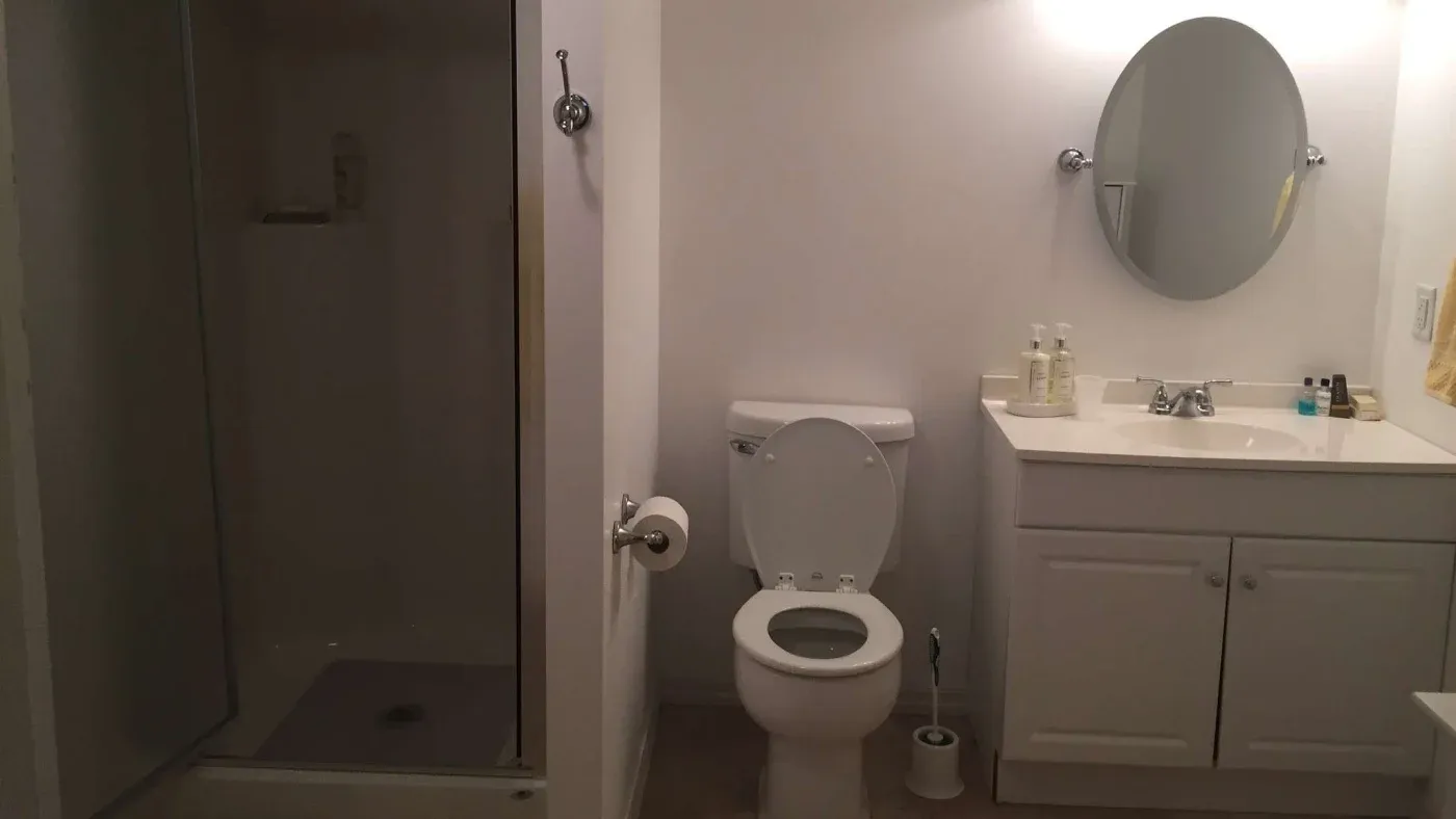 A well-lit bathroom with a shower on the left, central toilet, and a vanity on the right. Simple, clean design with a circular mirror above the sink.