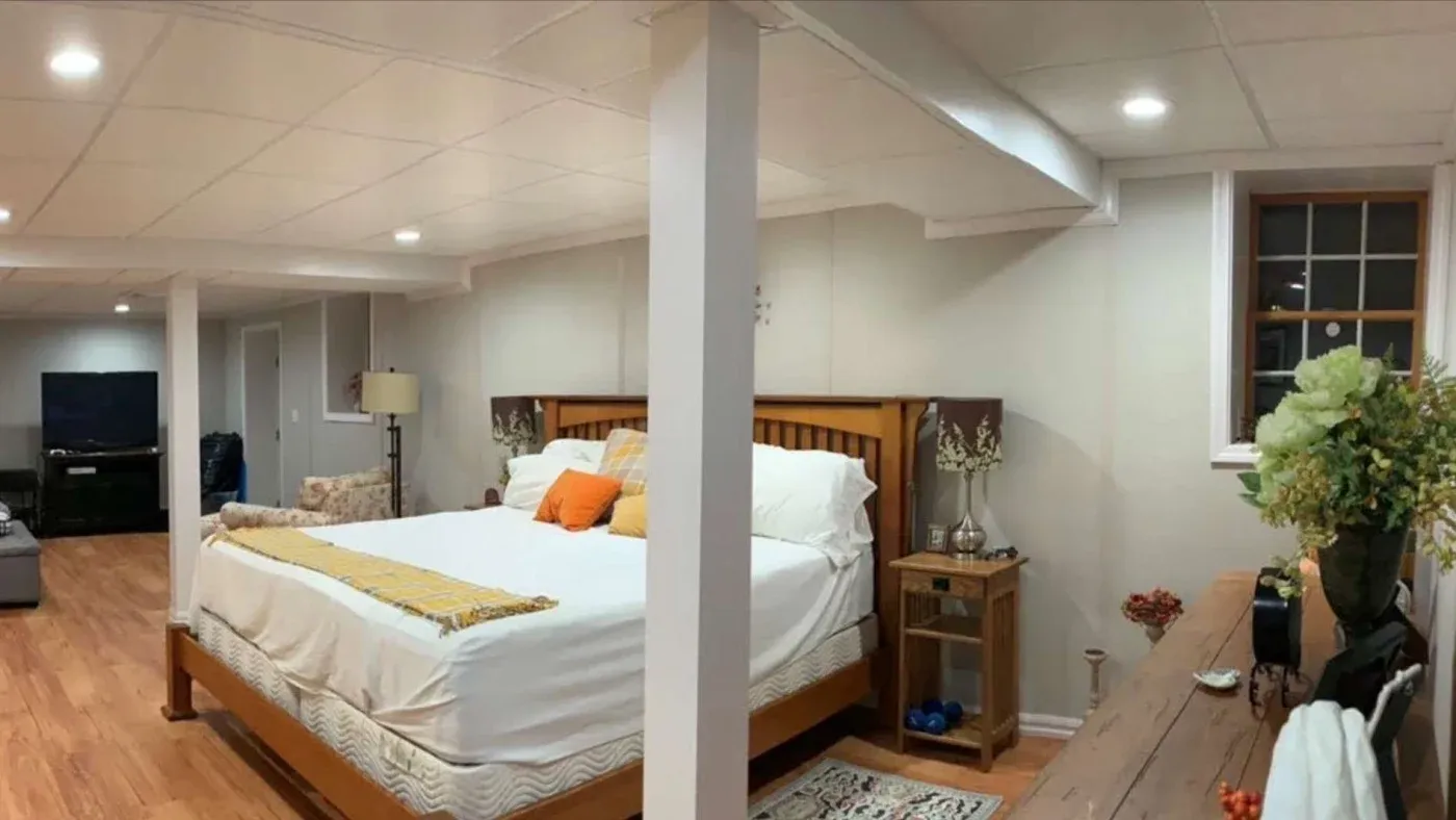 Spacious basement bedroom with a wooden bed, white bedding, and orange pillows. Warm lighting, wood floors, a bedside table with plants, and a large TV.