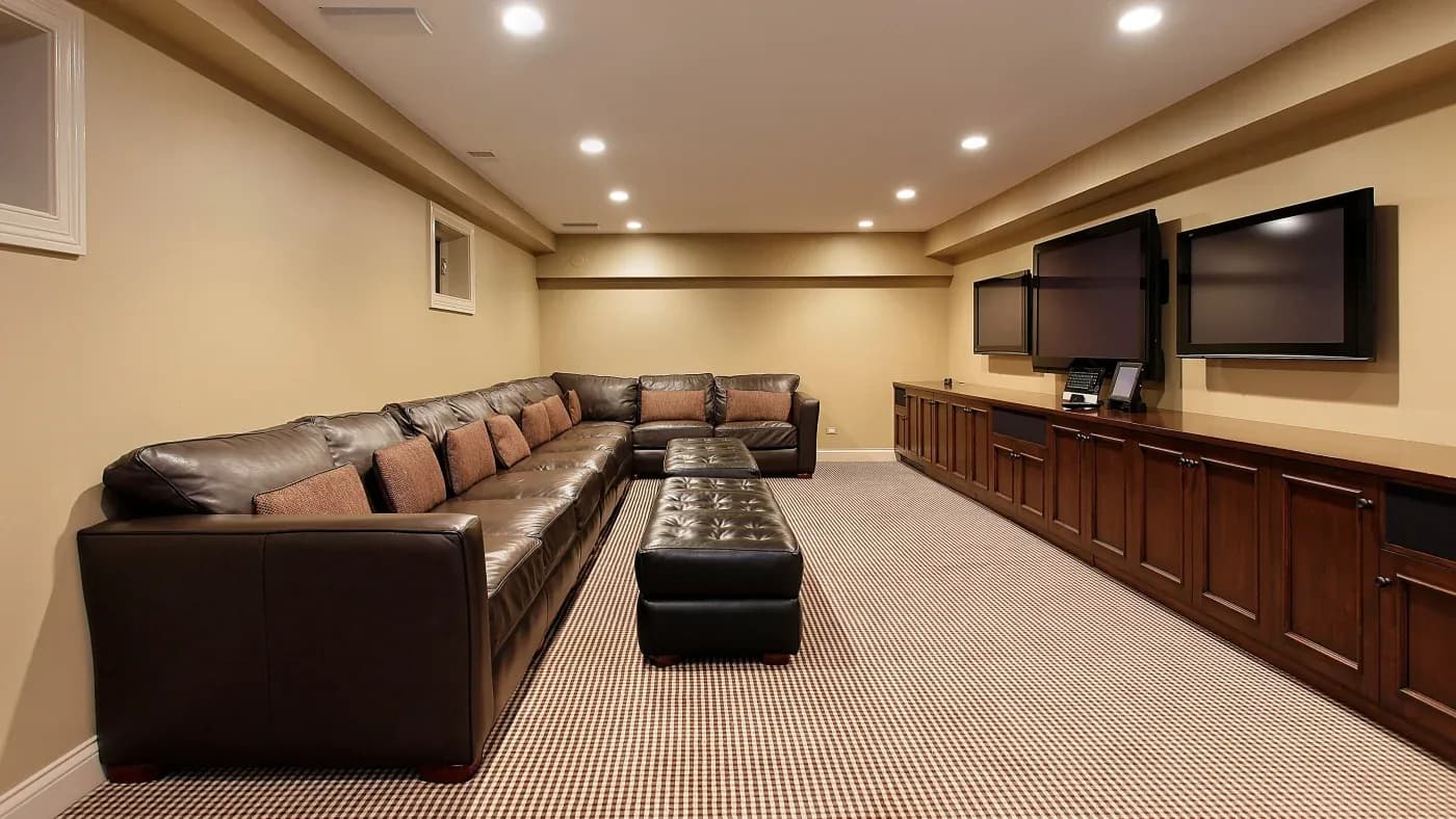 Spacious home theater room with a large brown leather sectional sofa and matching ottoman. Three flat-screen TVs are mounted on a wood-paneled wall.