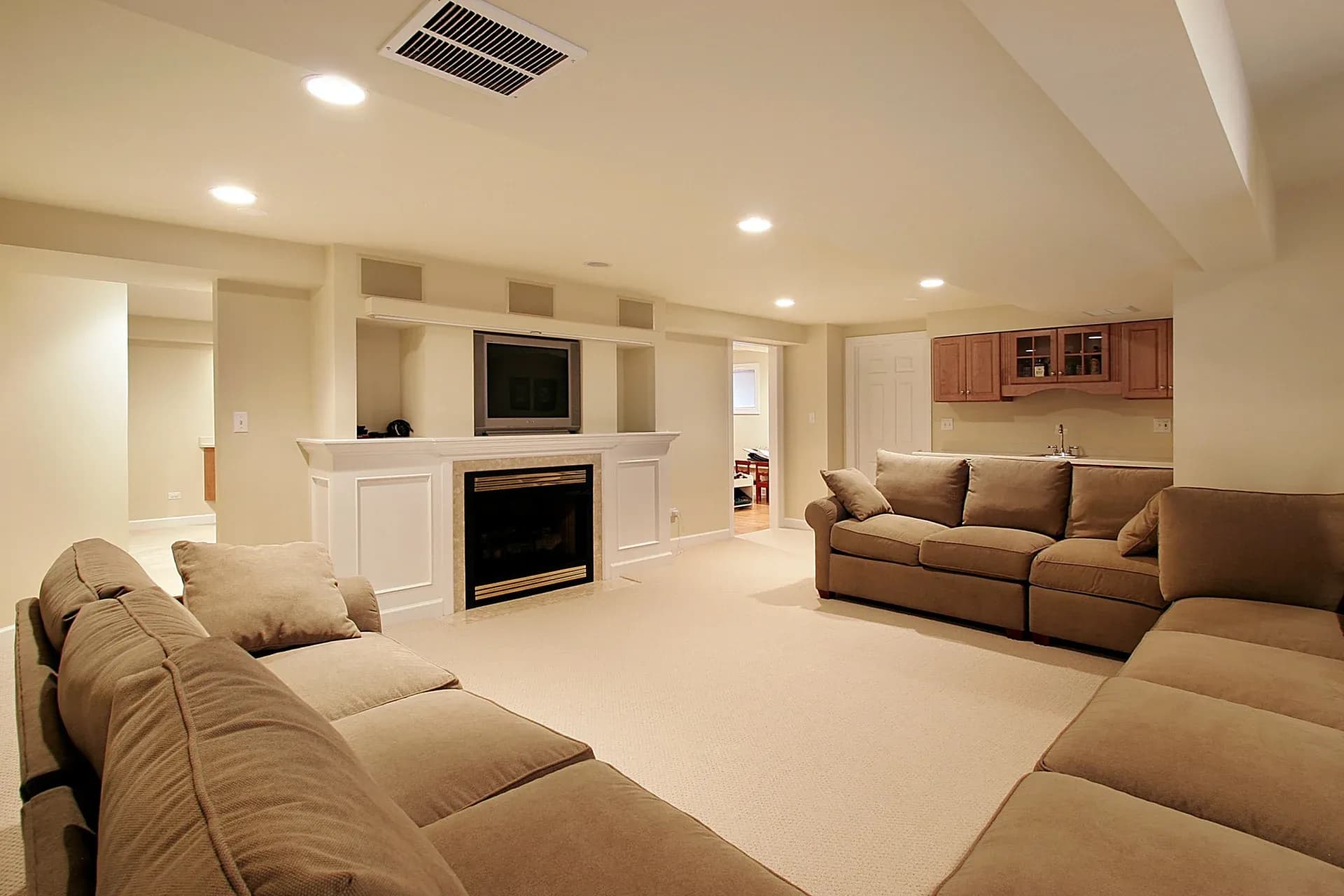 Spacious basement living room with beige walls, carpet, and dim lighting. Features cozy brown sofas, a TV above a fireplace, and a small kitchenette.