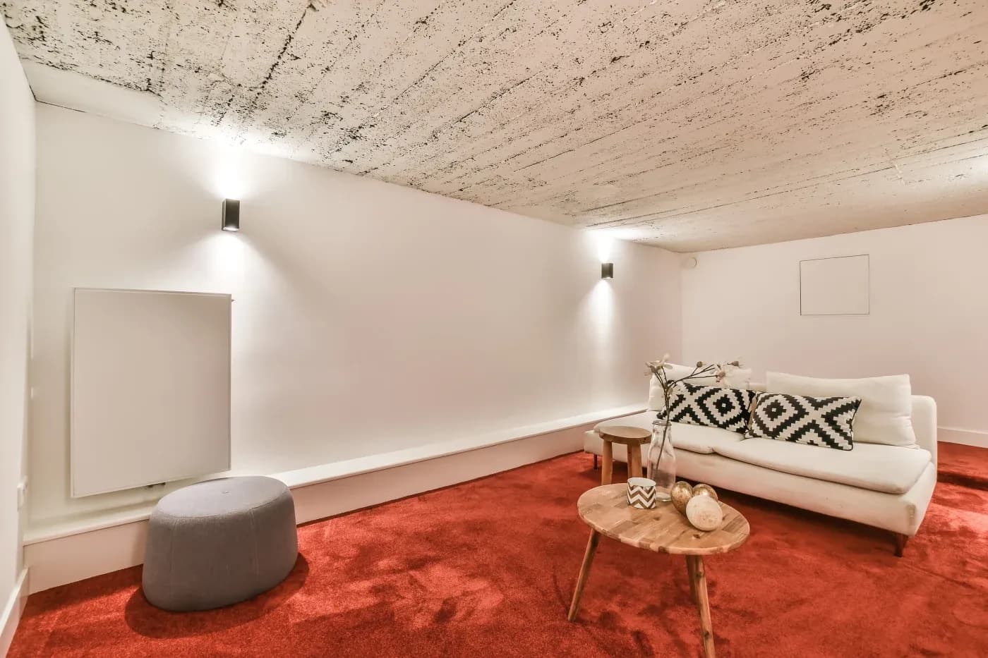 Modern room with a low ceiling, featuring a white sofa with geometric pillows, a wooden coffee table, and red carpet. Minimalist and cozy ambiance.