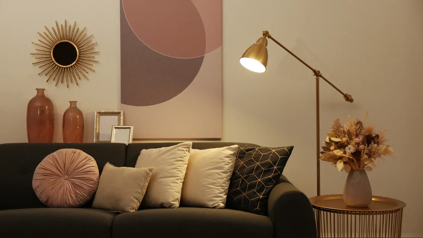 A cozy living room with a dark sofa adorned with soft pillows in pink, beige, and black. A modern lamp casts warm light on a side table with a vase and floral arrangement. Wall art and vases add elegance.