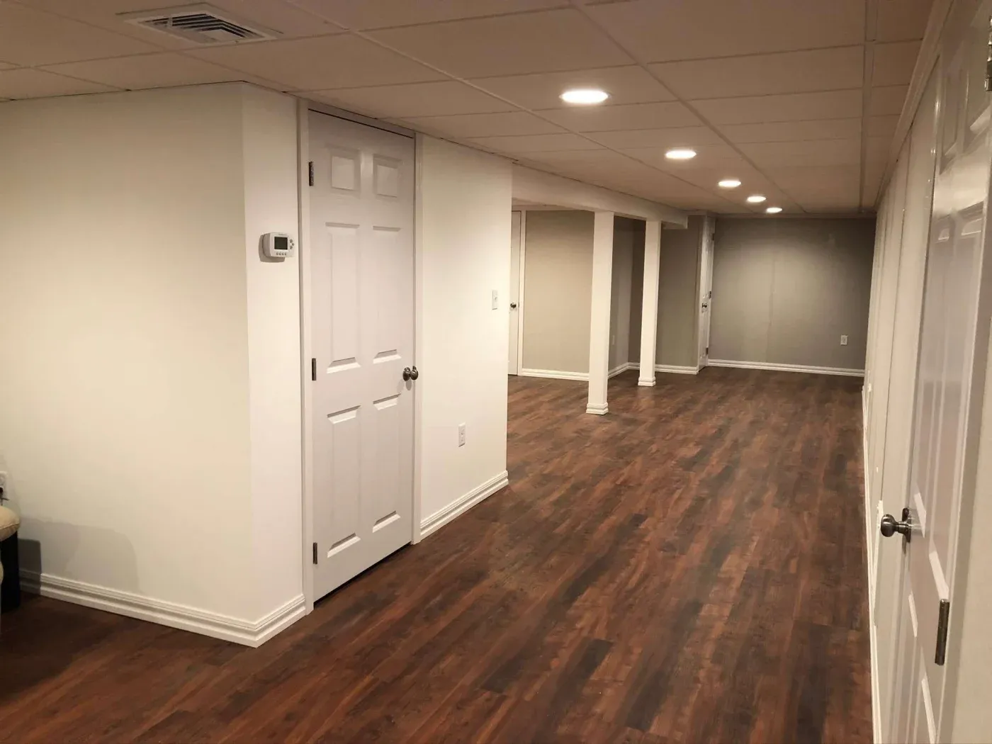Spacious finished basement with white walls, recessed lighting, and dark wooden floors. The room is elongated, featuring closed white doors.