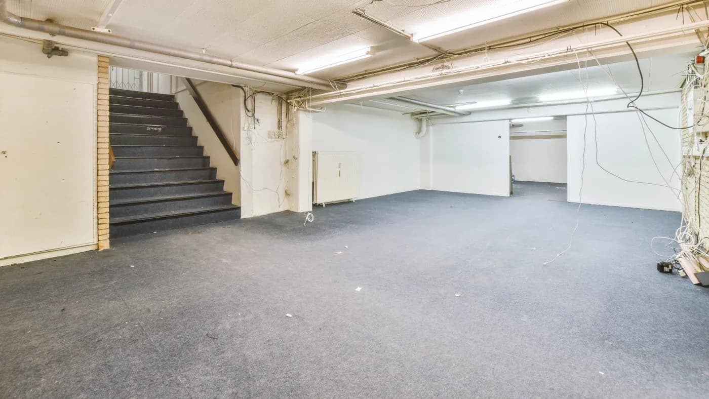 Spacious empty basement with plush gray carpet, exposed wires on walls, and a dimly lit atmosphere. Stairs lead up to the left; walls are plain white.