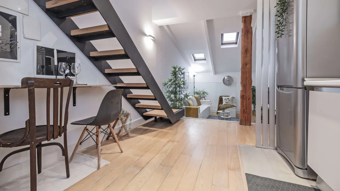 Modern interior of a loft apartment featuring a wooden staircase, cozy dining area with chairs, lush plants, skylights, and a bright, airy ambiance.