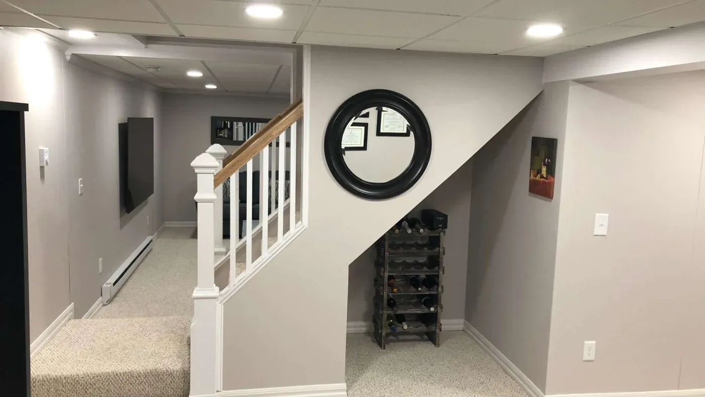 A cozy basement with light gray walls features a staircase with white railings. A round mirror hangs on the wall above a wine rack, adding a welcoming feel.