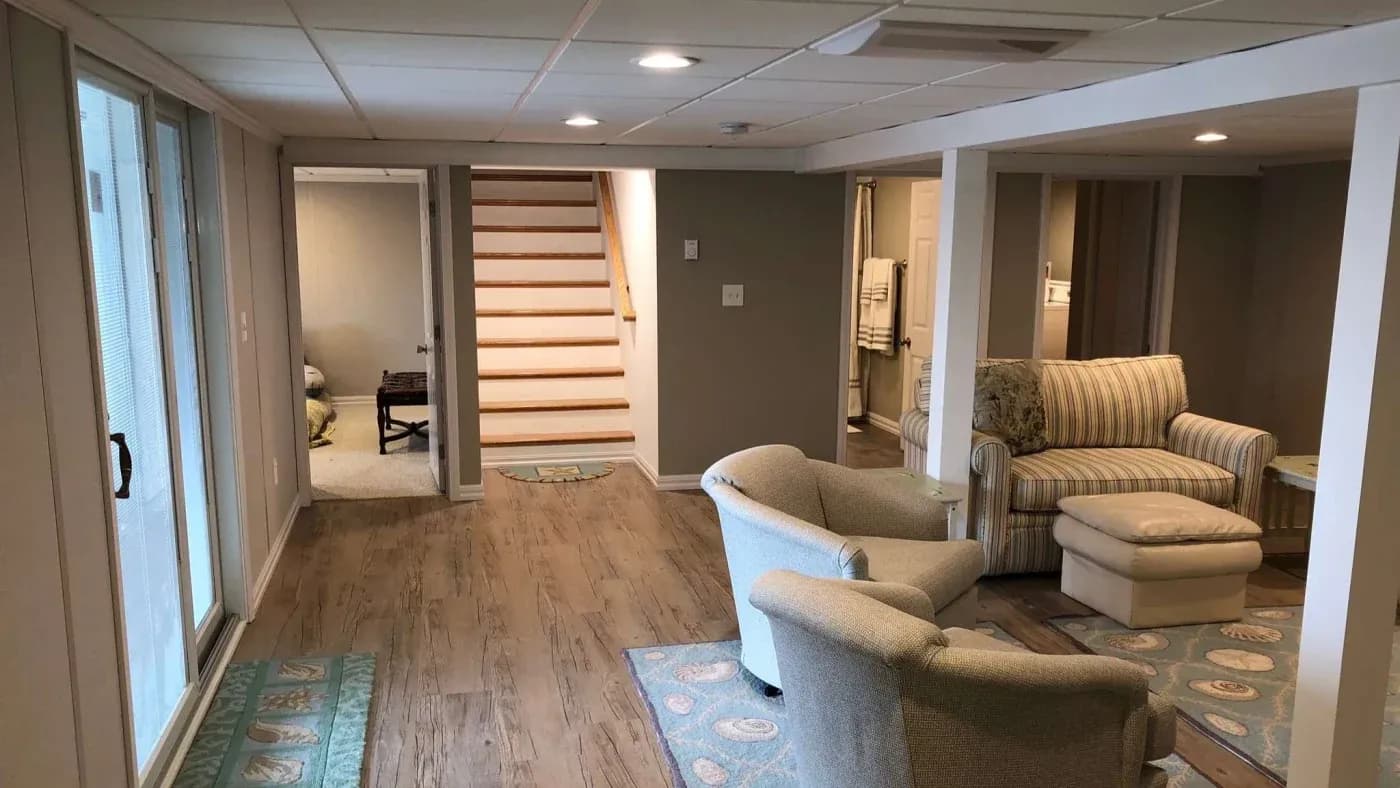 A cozy basement with a seating area featuring armchairs and a striped sofa. Wooden stairs lead upstairs, and sliding glass doors let in natural light.
