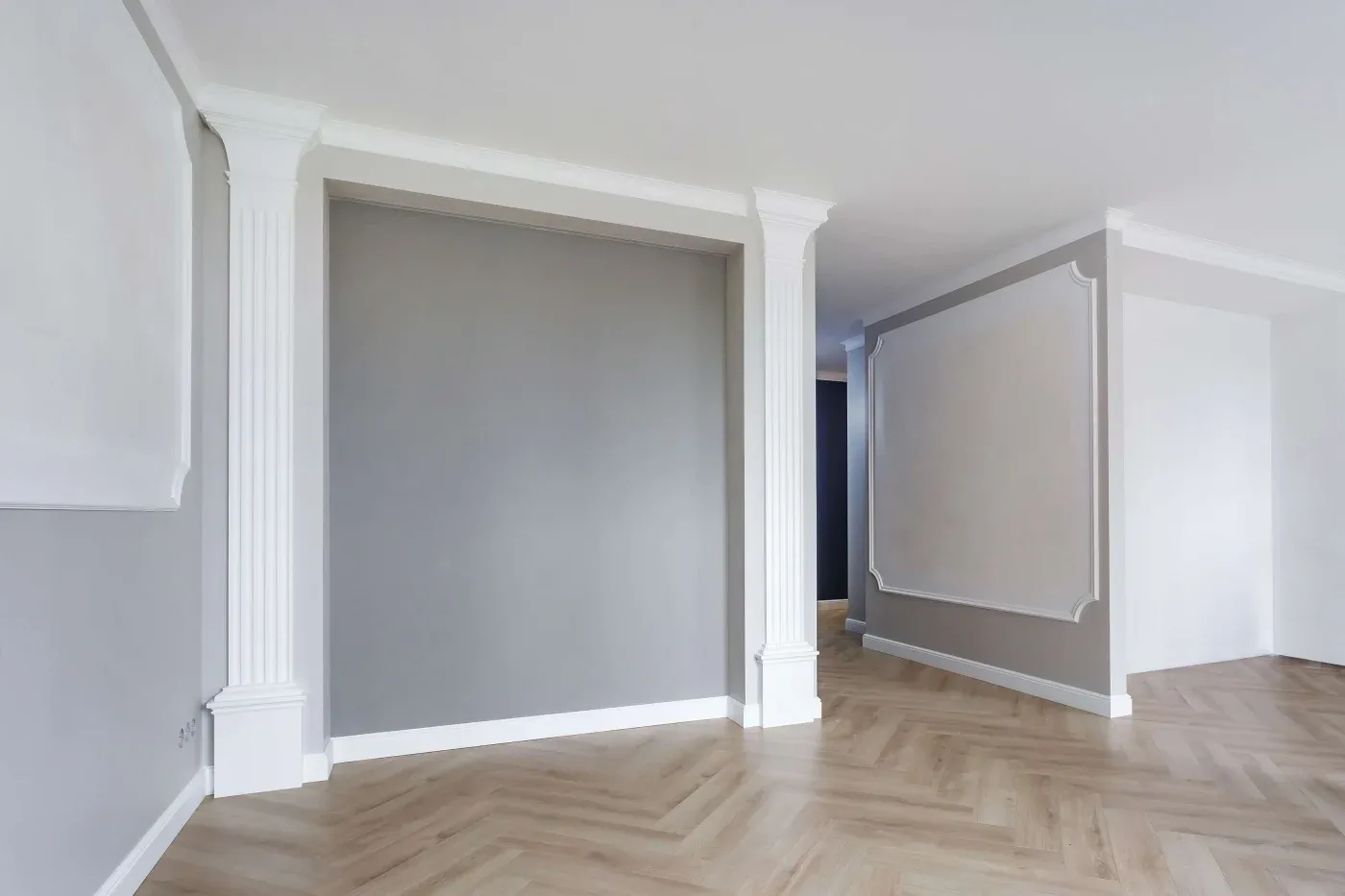 A modern, empty room with light gray walls, white decorative columns, and a chevron-patterned wooden floor, conveying a clean, elegant atmosphere.
