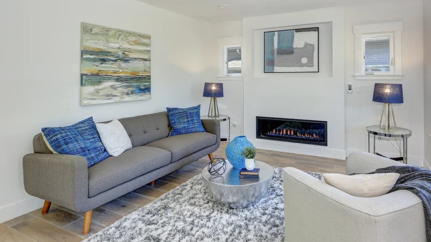 A modern living room features a gray sofa with blue plaid cushions, a soft armchair, abstract art, a sleek fireplace, and a cozy atmosphere.
