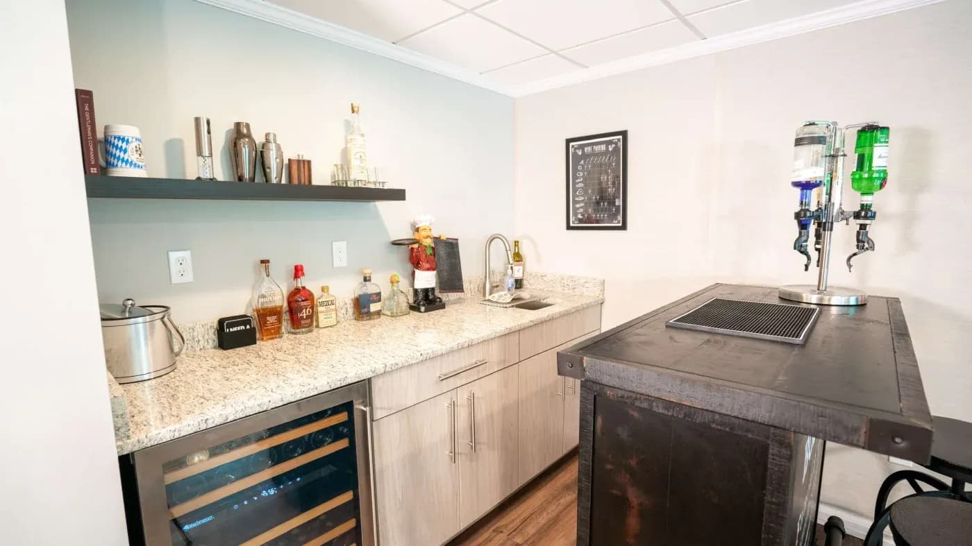 A modern home bar with granite countertops, bottles, a small sink, and a beer tap. Cozy ambiance with a wine fridge and a framed print on the wall.
