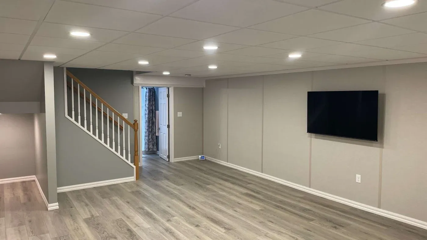 A modern basement with light gray walls and wood flooring. Features a wall-mounted TV, recessed lighting, and a staircase with wooden railing. A door at the back adds depth.