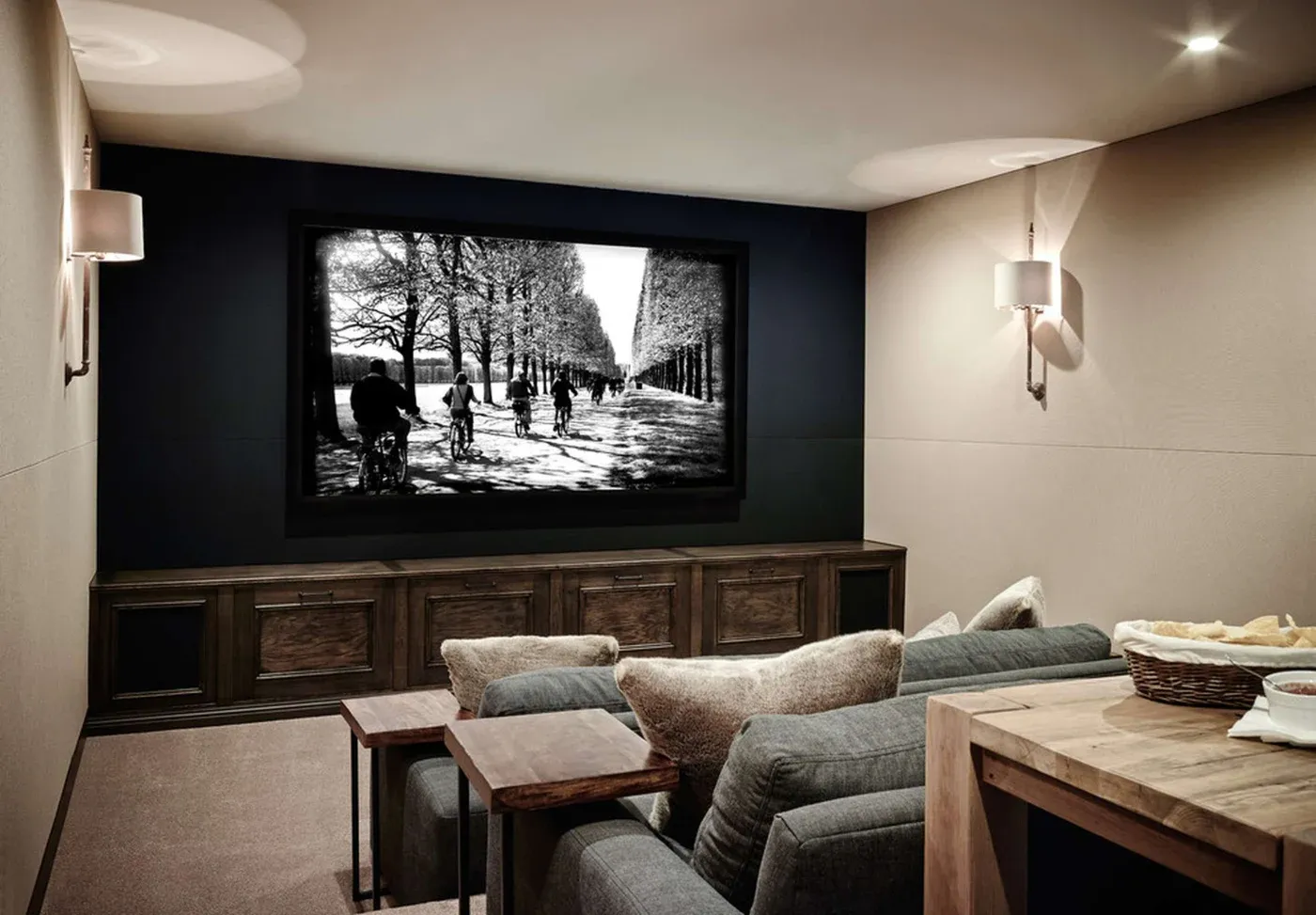 Cozy home theater with plush gray couches and a large wall-mounted screen displaying a black-and-white park scene. Warm lighting adds comfort.