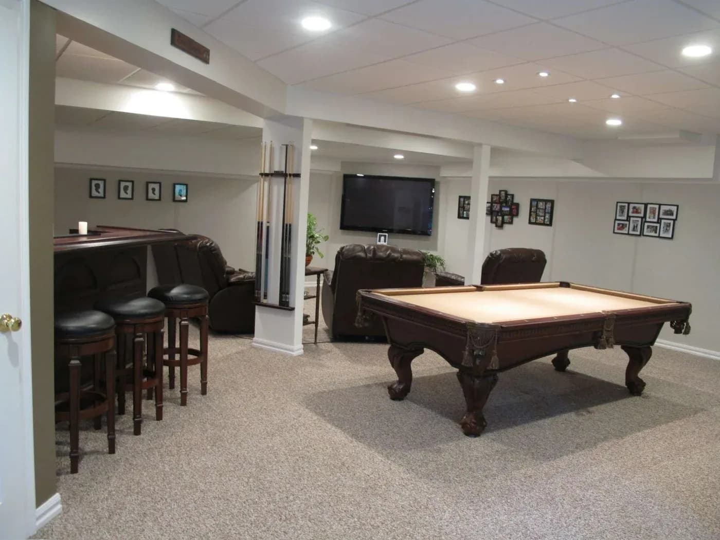 A cozy basement game room with a pool table, wall-mounted TV, and leather recliners. Framed photos and a small bar area add a warm, inviting feel.