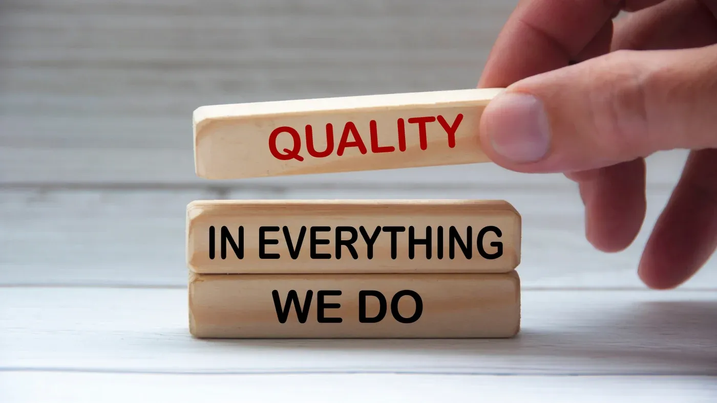 A hand places a wooden block with "QUALITY" in red text on two blocks reading "IN EVERYTHING WE DO" in black, conveying a commitment to excellence.