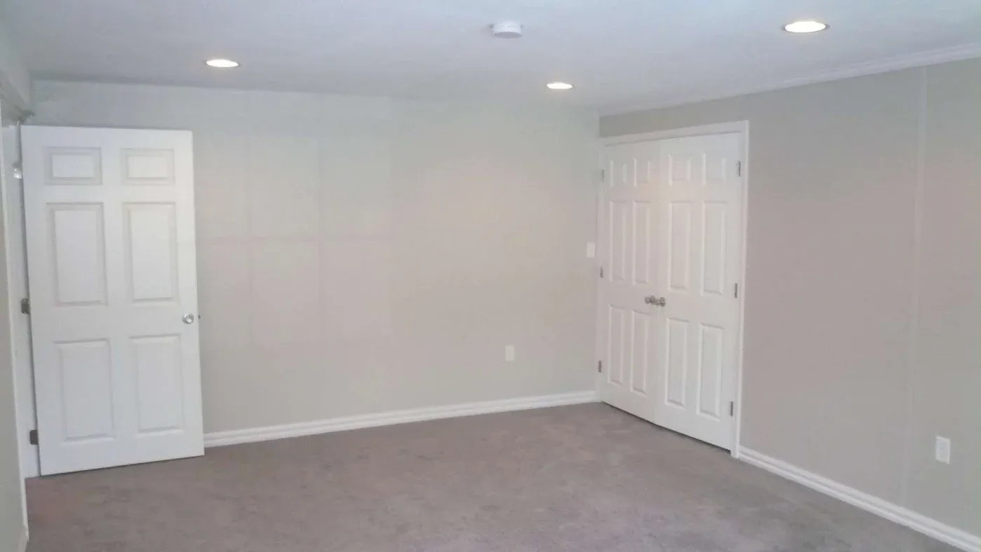 A clean, empty room with beige walls and carpet. It features a white door on the left and double doors on the right. Recessed ceiling lights emit a soft glow.