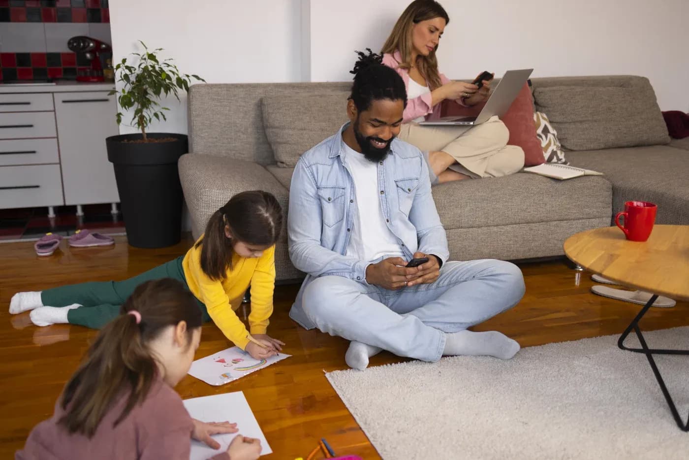 A family in a cozy living room, two children drawing on the floor, a man smiling at his phone, and a woman working on her laptop on the couch.