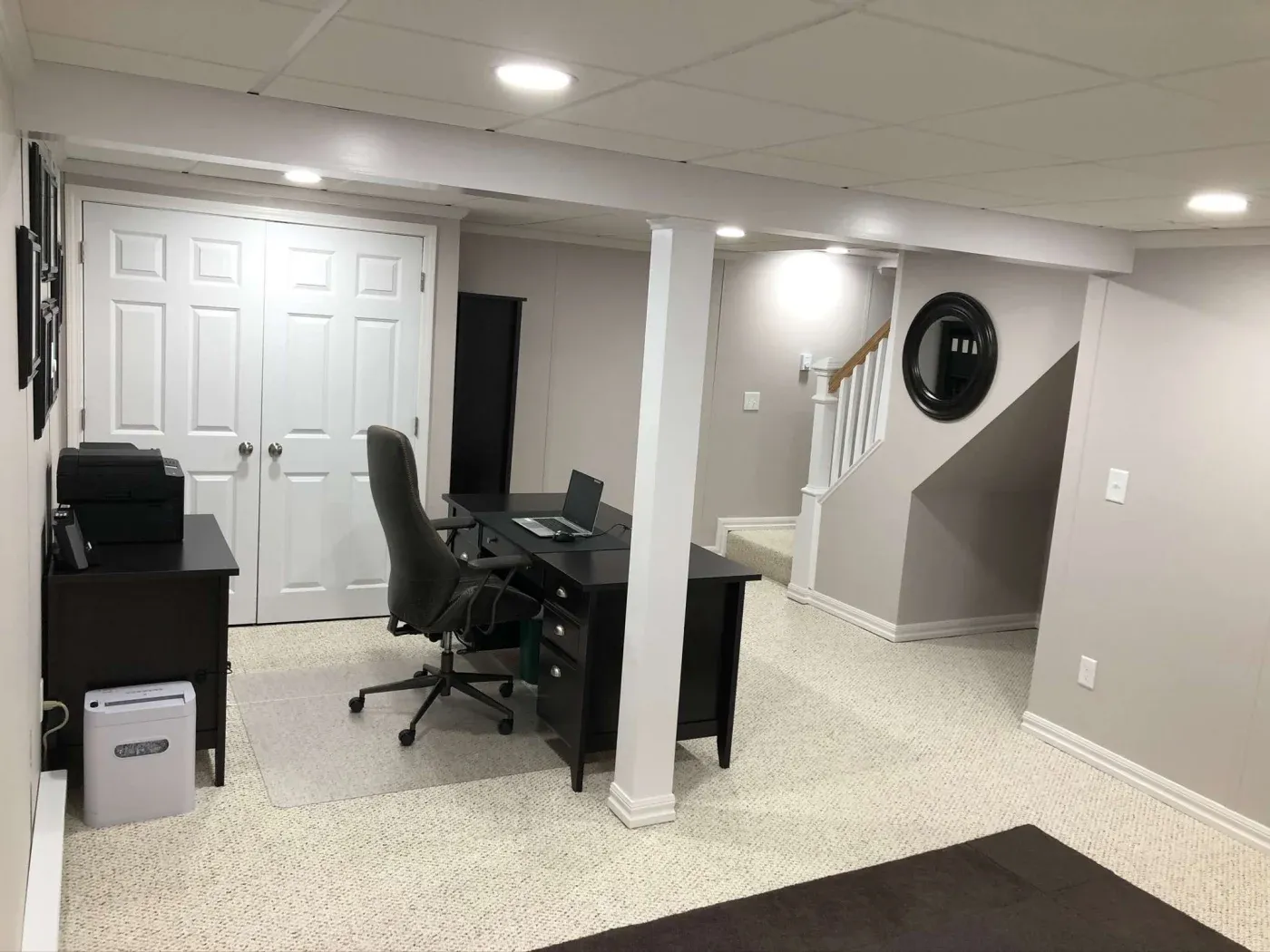 A modern, organized home office with a black desk, swivel chair, and laptop. Neutral tones, soft lighting, and a round mirror add warmth.