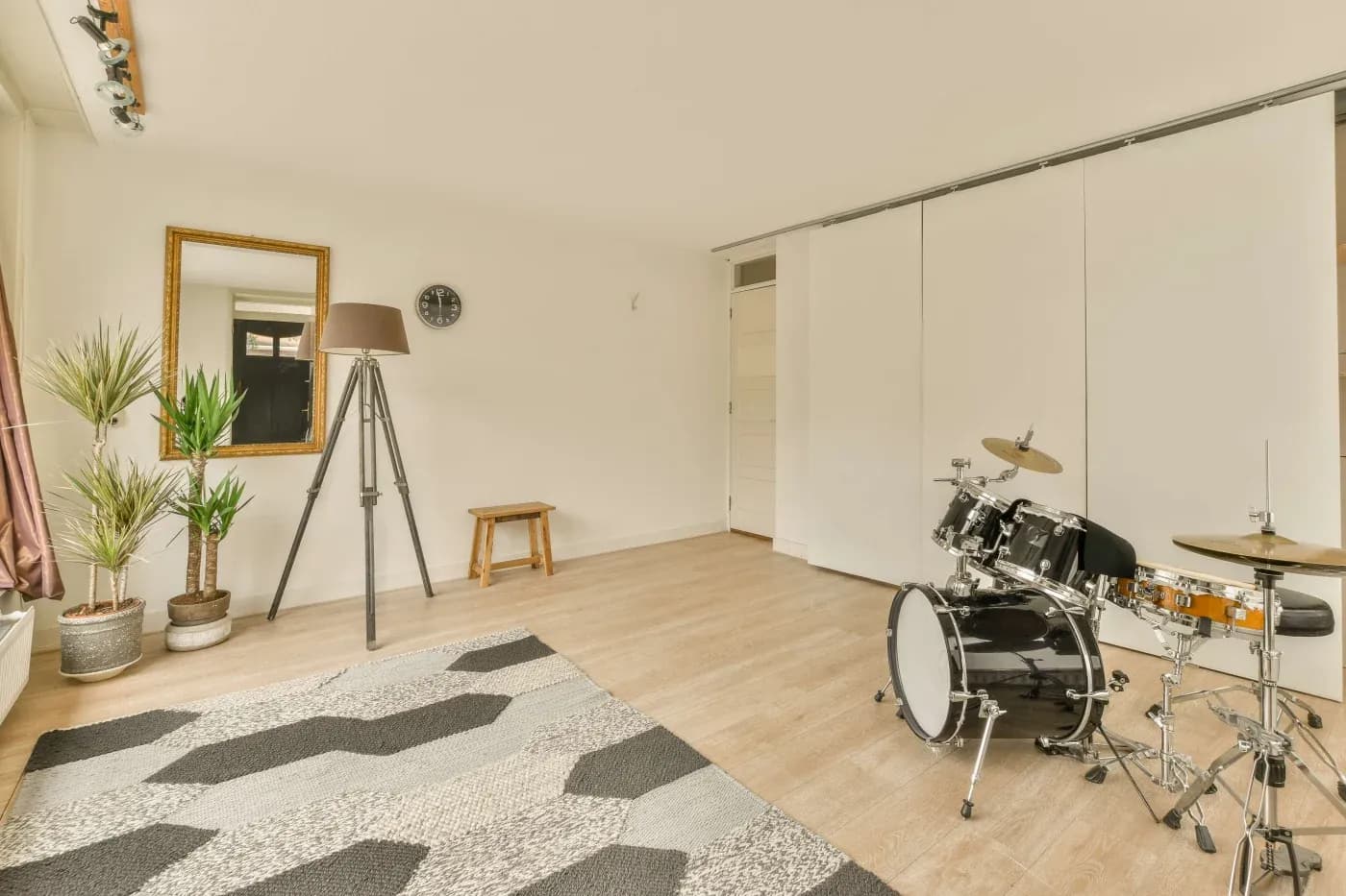 Minimalist room with a drum set, tripod lamp, mirror, and potted plants on a wooden floor. A geometric rug adds a modern touch. Bright and open.