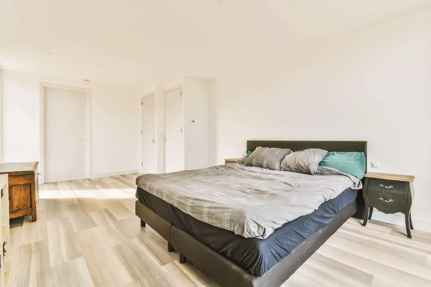 A bright, minimalist bedroom with a neatly made bed centered. Light wooden floors, white walls, and simple furniture create an airy, calm ambiance.