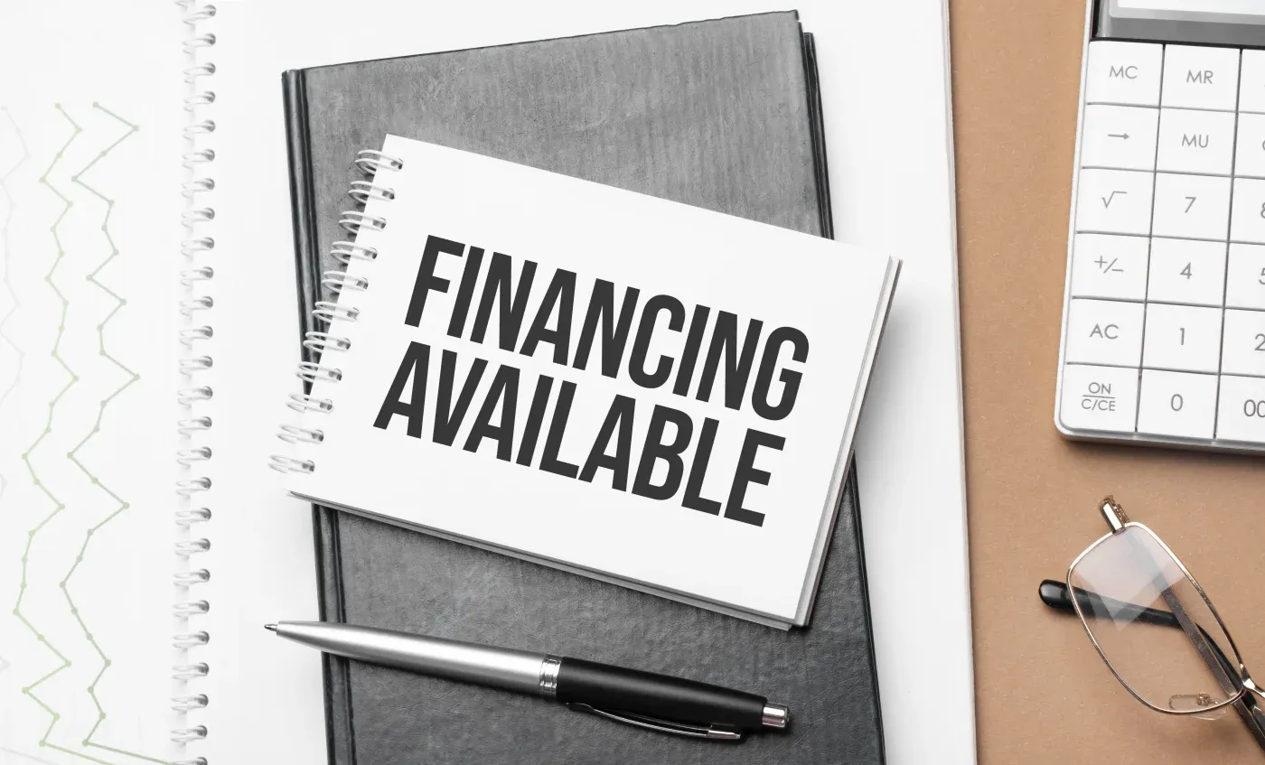 A notebook open on a desk displays the text "FINANCING AVAILABLE" in bold. Nearby are a pen, eyeglasses, graph paper, and a calculator, suggesting financial planning.