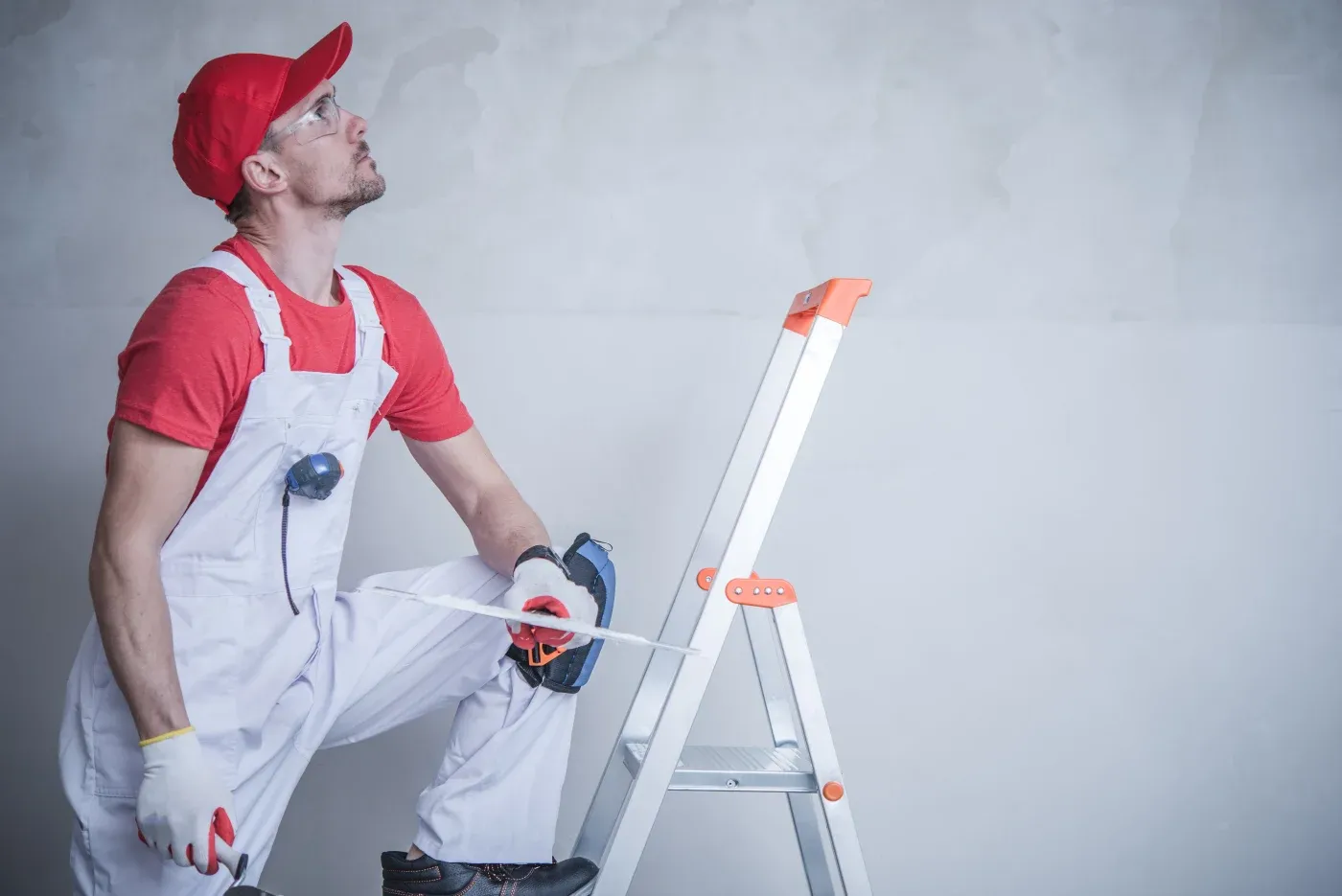 Man in red hat and overalls stands on a ladder, holding a power drill and measuring tape, looking upwards. The scene conveys focus and determination.