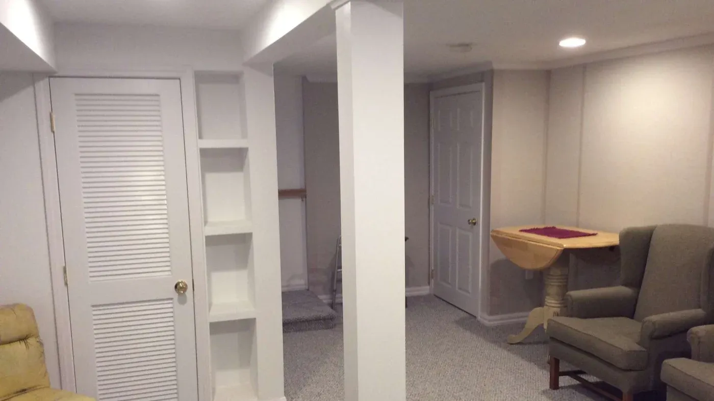 A cozy basement with a white door and open shelves on the left. Beige walls, armchairs, and a small wooden table create a warm, inviting atmosphere.
