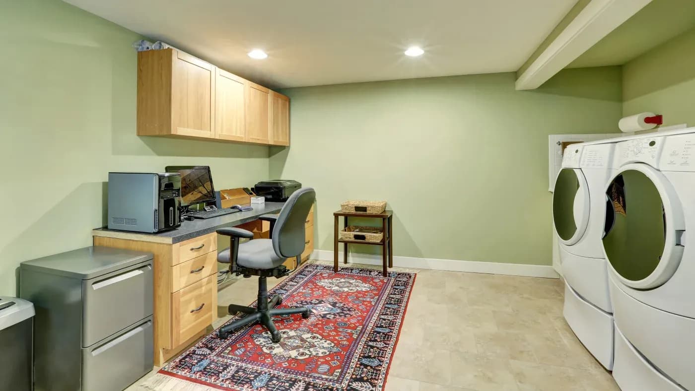 Laundry room with green walls, featuring a modern washer and dryer, a wooden desk with office chair, and a vibrant red patterned rug on tiled floor.