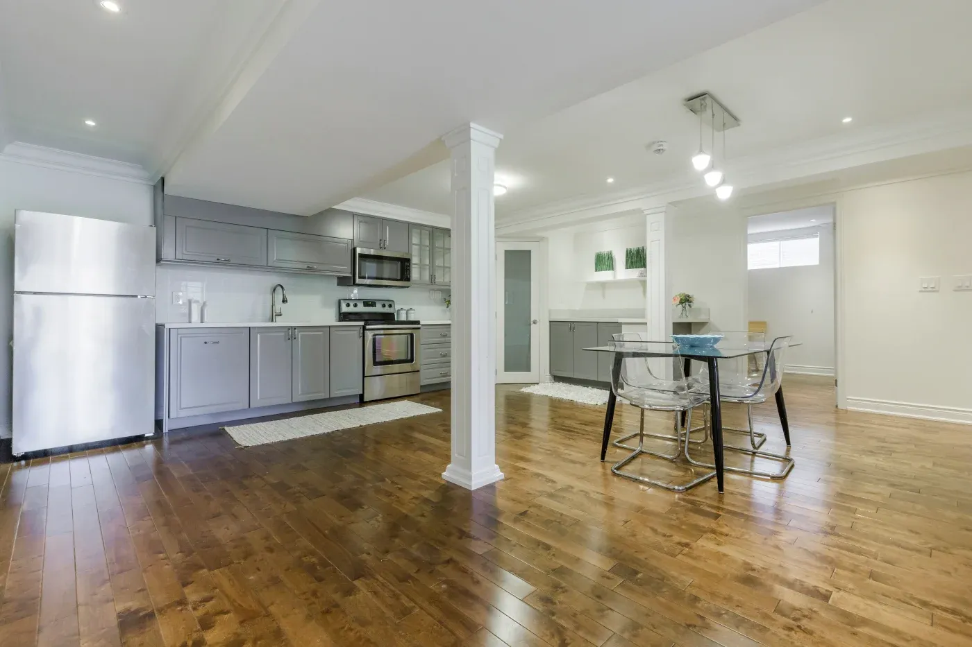 Modern kitchen with gray cabinets, stainless steel appliances, and wooden flooring. Adjacent dining area features a glass table and transparent chairs under stylish lighting.