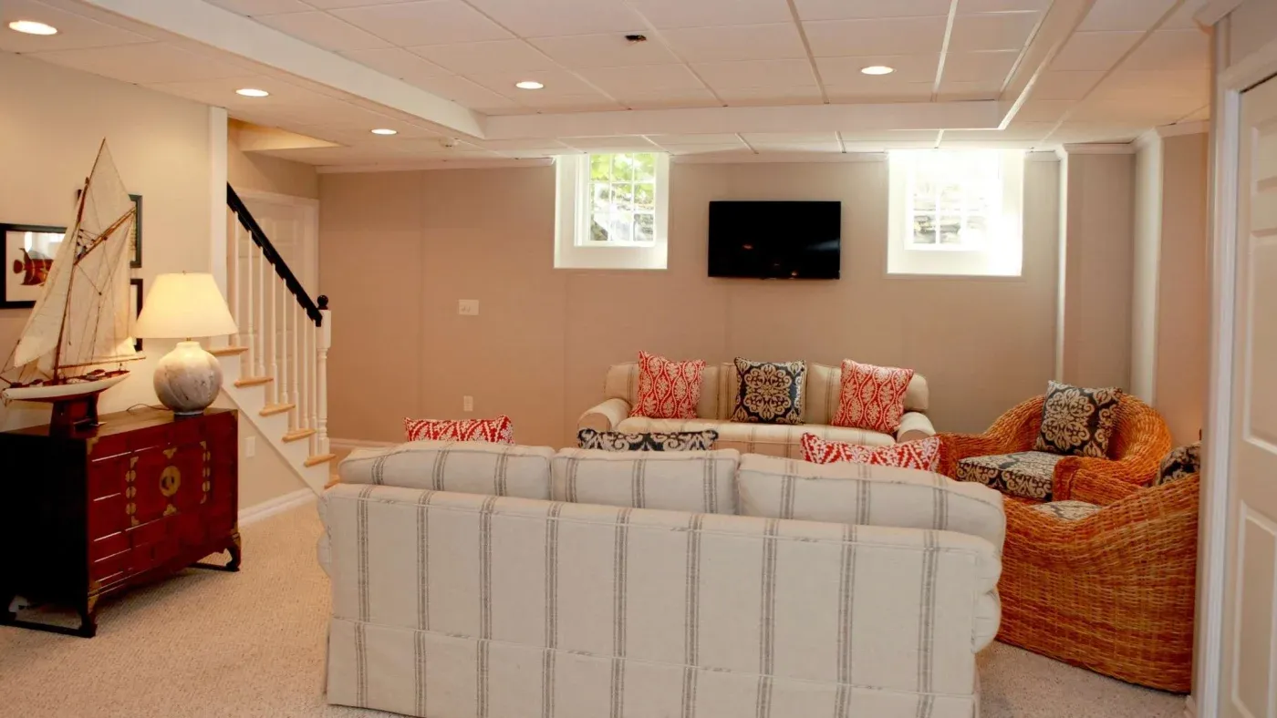 Cozy basement living room with beige walls, light sofas, and colorful patterned cushions. A wicker chair, TV, and decorative ship create a welcoming feel.
