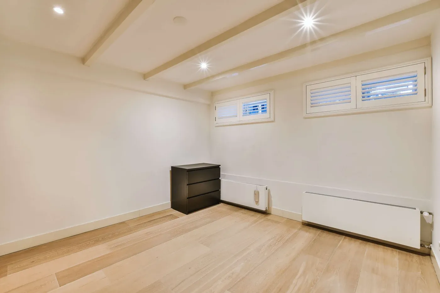 A minimalistic room with light wood flooring, off-white walls, and two small windows. A black dresser and radiators add contrast, creating a serene atmosphere.