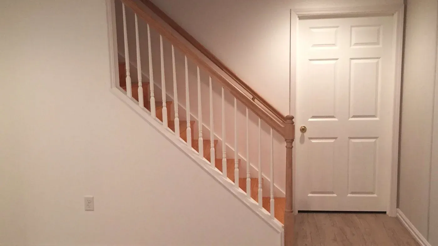 A staircase with wooden handrail and white balusters leading up, next to a closed white door on a neutral-toned wall, conveys simplicity and calm.